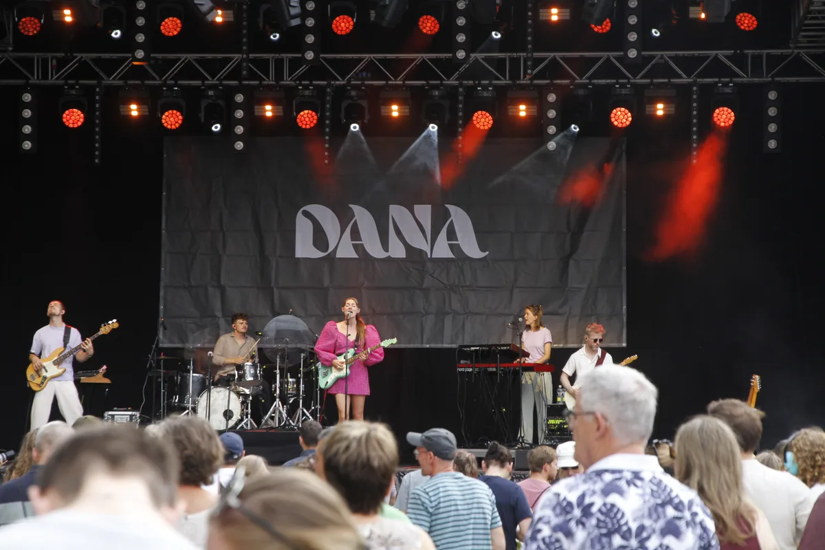 Dana und ihre Band spielen auf einer schwarzen Bühne. Oben leuchten rote Scheinwerfer. Die Sängerin trägt ein rosa Kleid und spielt auf einer grünen Gitarre.